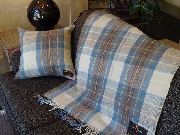 The Stewart Muted Blue Deluxe Highland Blanket, featuring blue, brown, and cream plaid, is draped over a dark gray sofa with a matching pillow. A lamp and plant in the background enhance the cozy atmosphere.