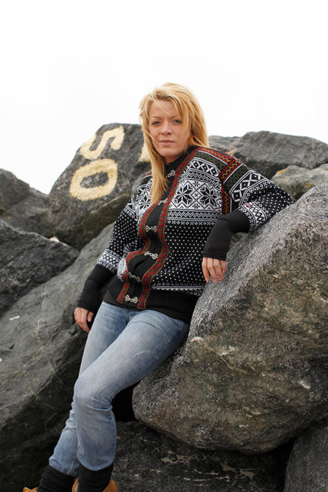A person wearing a Nordlys of Norway Northern Lights patterned cardigan in charcoal grey and natural leans against large rocks, one of which has the number 50 painted on it, under an overcast sky.
