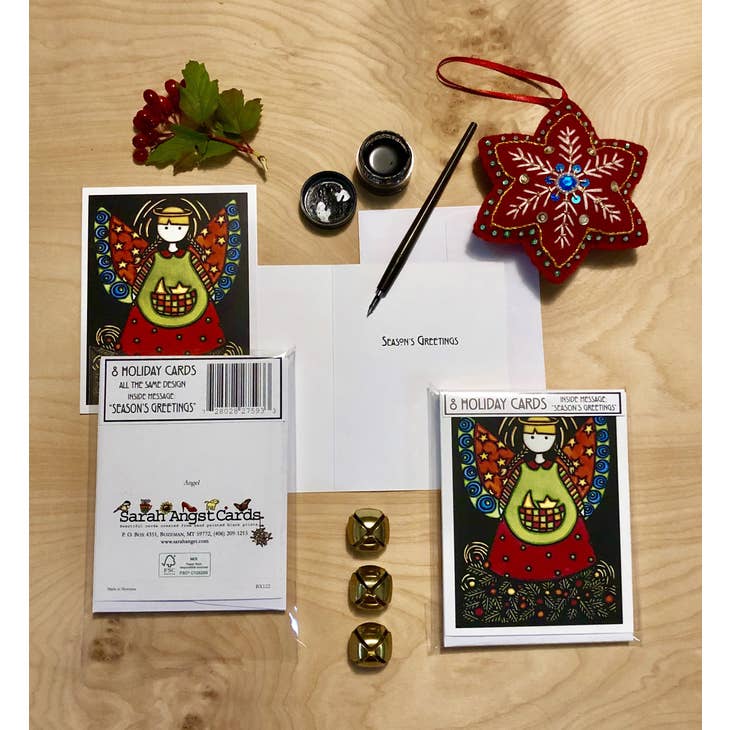 The "Angel Packaged 8 Holiday Cards" by Sarah Angst Art showcases a collection of holiday greeting cards featuring an angel design, a pen, bells, a festive ornament, and a small branch with berries on a wooden surface. Each card is crafted from FSC Paper and includes envelopes with the message "Season's Greetings.