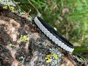 The Traditional Sami Single Braid Bracelet - Black #1, handcrafted in Lapland, Sweden from reindeer leather with a silver braid, rests on tree bark amid green grass and yellow lichen.