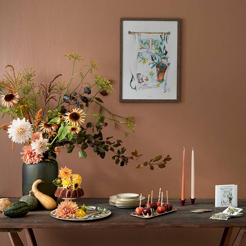 A table adorned with autumn-themed decor, showcasing a bouquet of flowers, plates, candles, caramel apples, and artwork in a warm-toned room. The "Room with a View" card pack, featuring a square card folder with eight cards and eucalyptus envelopes, adds an elegant touch to this cozy setting.