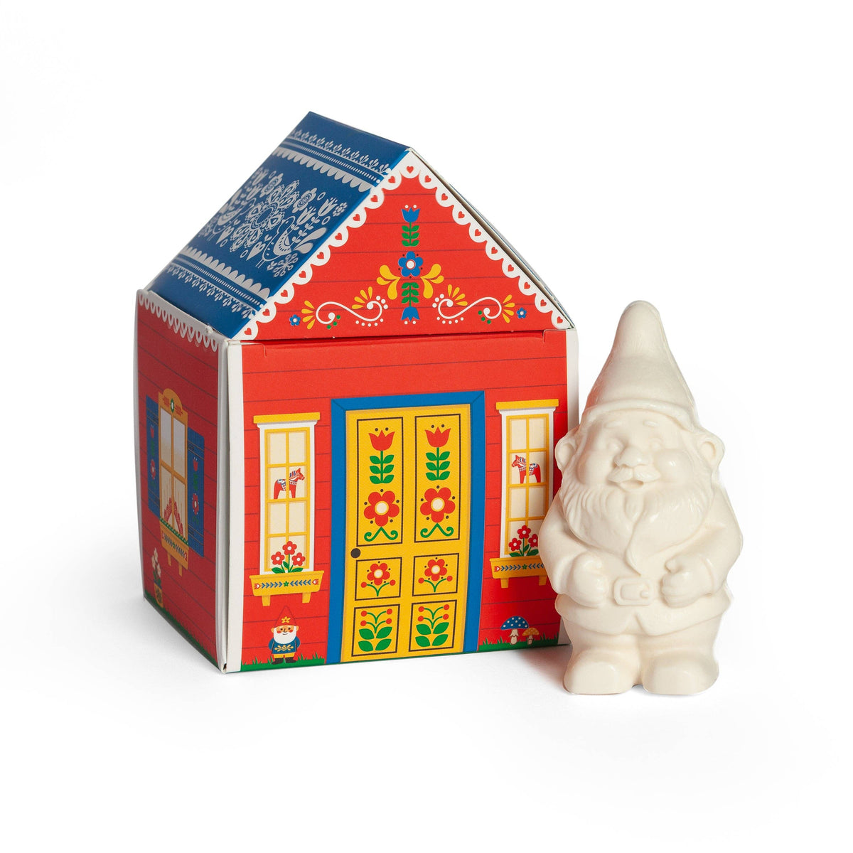 A small white gnome figurine next to a red house-shaped box with Nordic folk art patterns, containing bar soap labeled “Gnome’s Cottage.”.