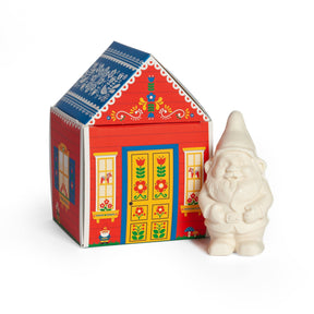 A small white gnome figurine next to a red house-shaped box with Nordic folk art patterns, containing bar soap labeled “Gnome’s Cottage.”.