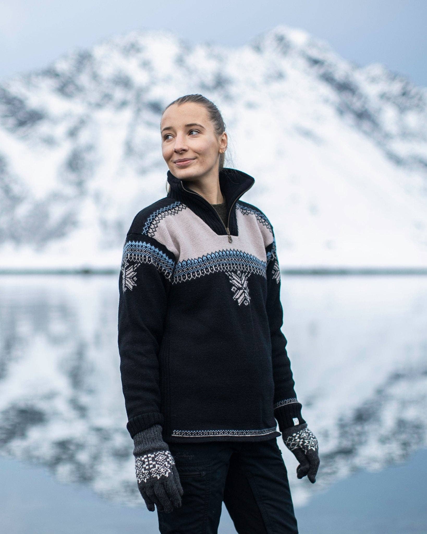A woman wearing a Voss Women's Sweater (Black) stands outdoors in front of a snowy mountain and a body of water.