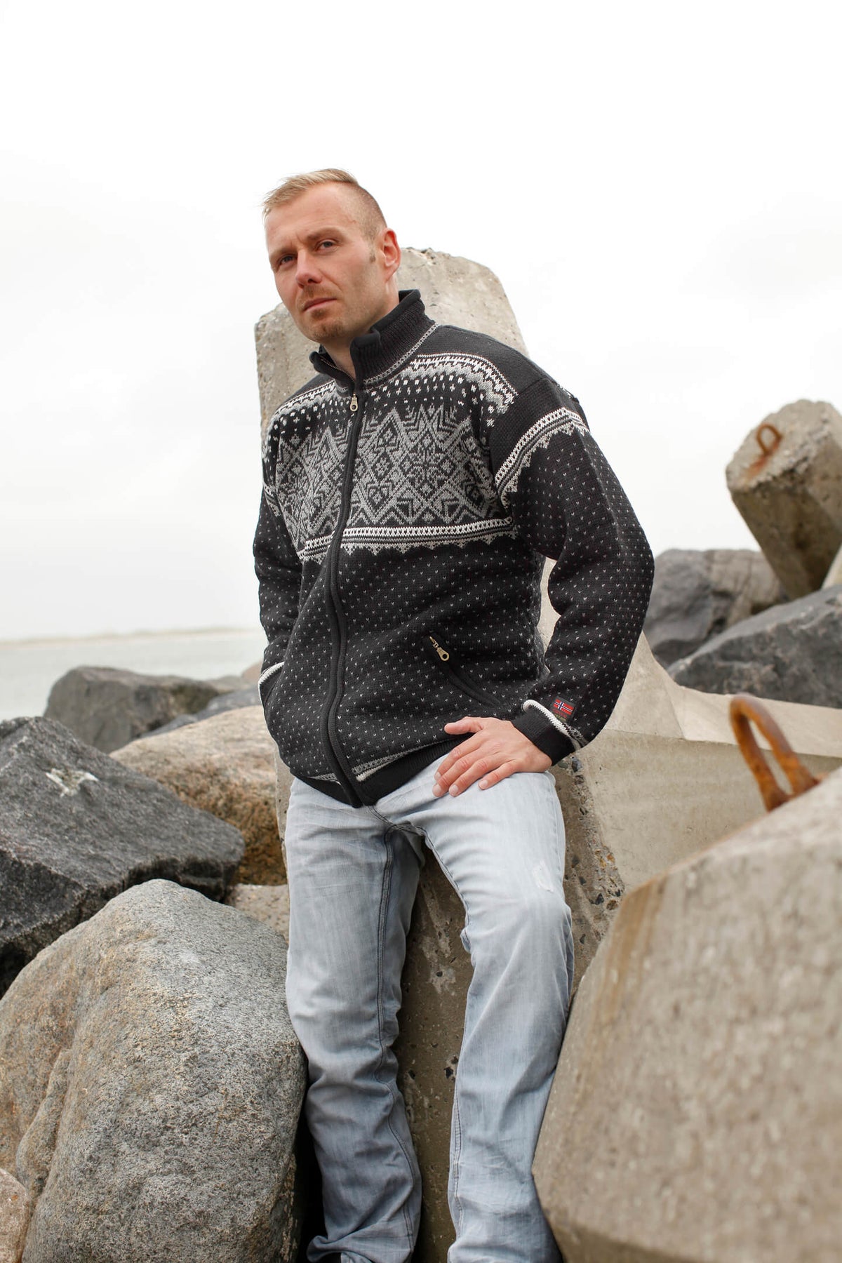 A man wears a black zip-up Norwegian wool jacket featuring a white snowflake and geometric pattern across the chest and shoulders.