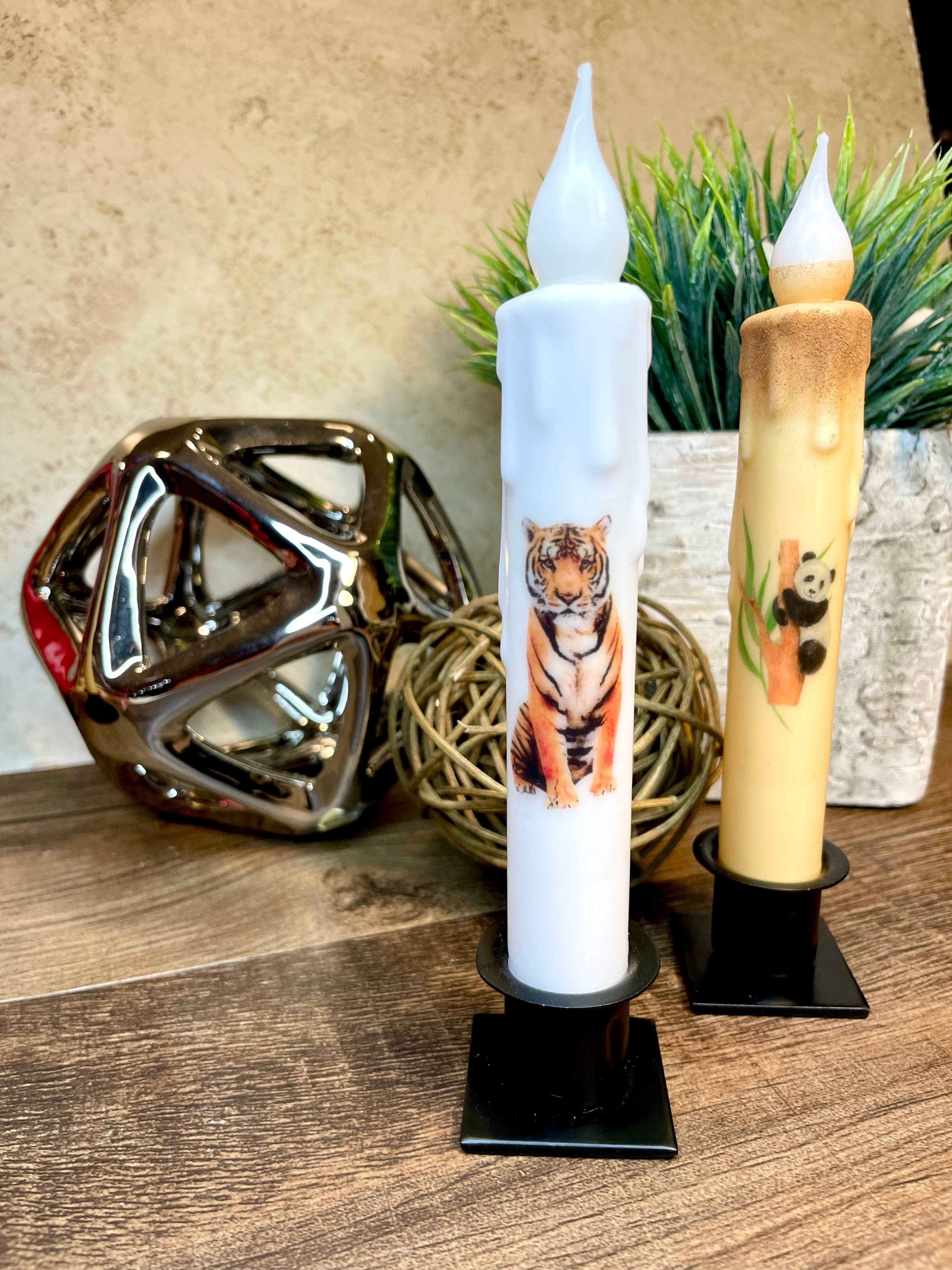 Two decorative candles featuring tiger and panda designs are elegantly placed on a wooden surface, each held by a Square Wrought Iron Taper Holder. Behind them, there's a wicker ball, a metallic geometric sculpture, and a potted plant, creating a harmonious blend of rustic and modern elements.