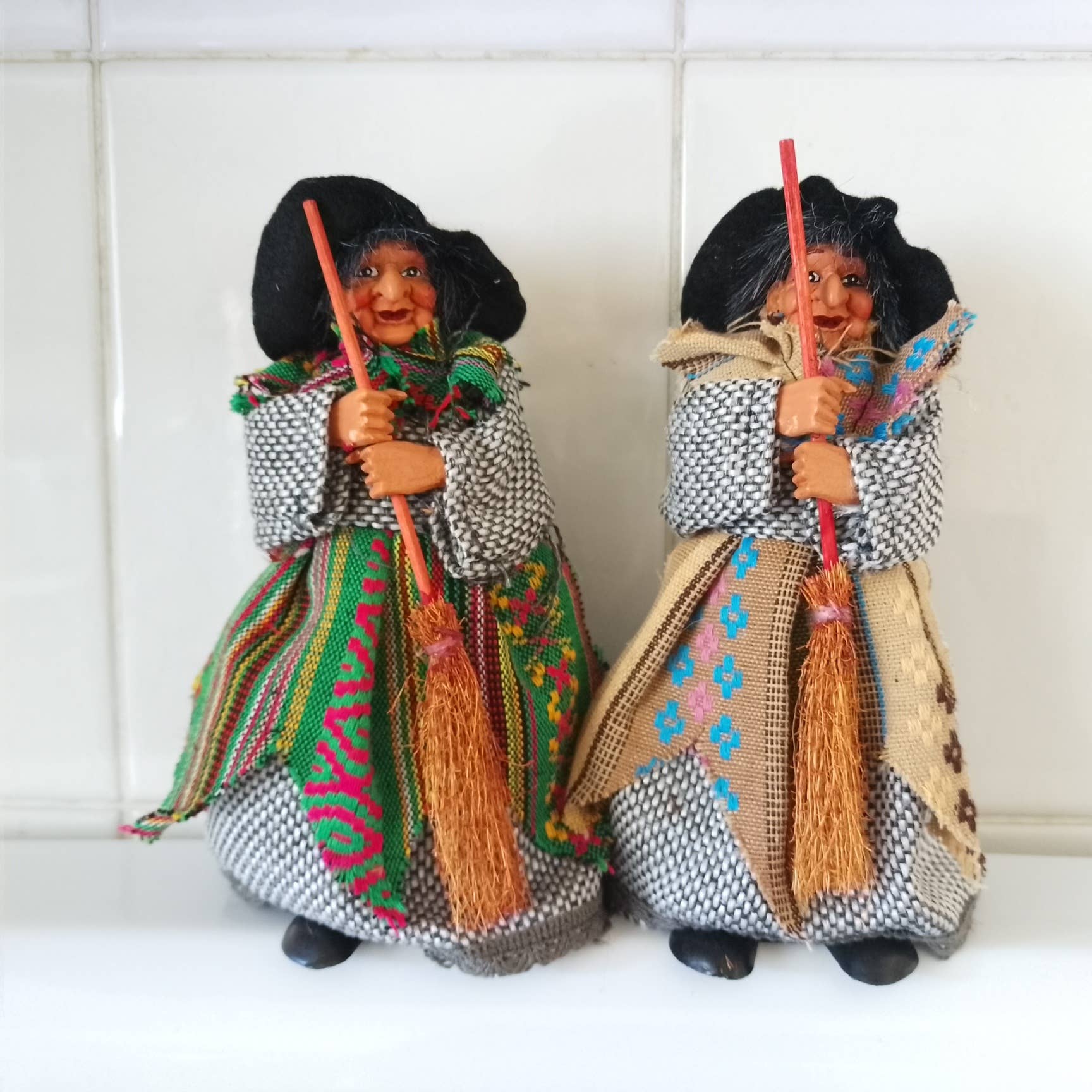Two Yasmina Green Kitchen Witch figurines, each 7" tall and dressed in patterned shawls and long dresses, stand side by side with brooms against a tiled background.