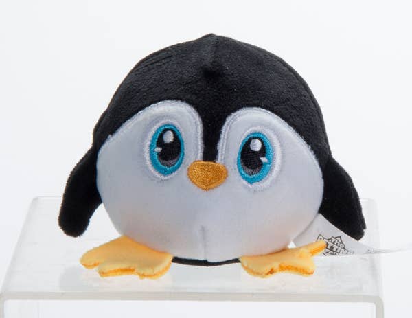 The Plush: Squishiez Penguin Small 3", featuring blue eyes, an orange beak, and flippers, sits adorably on a clear surface, adding a touch of squishable fun to your plush toys collection.