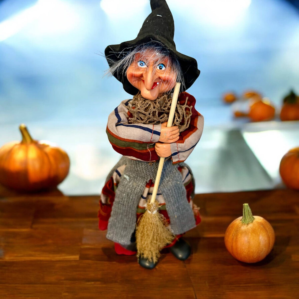 The Marketplace24 Kitchen Witch: Nonna Red 10" Tall with Broom stands on wood among pumpkins, bringing Scandinavian good luck charm to your Nordic kitchen decor. Her colorful dress and black hat pop against the blue background for a festive touch.