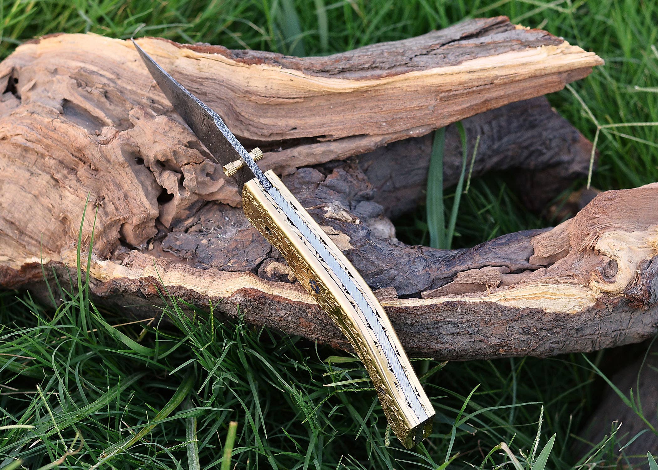 An Exquisite Golden Folding Knife with a brass handle and stainless steel blade rests on weathered wood, surrounded by grass.