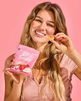 Woman in a pink shirt holds a pink bag labeled Swedish candy and displays a single candy piece, with a plain pink background.