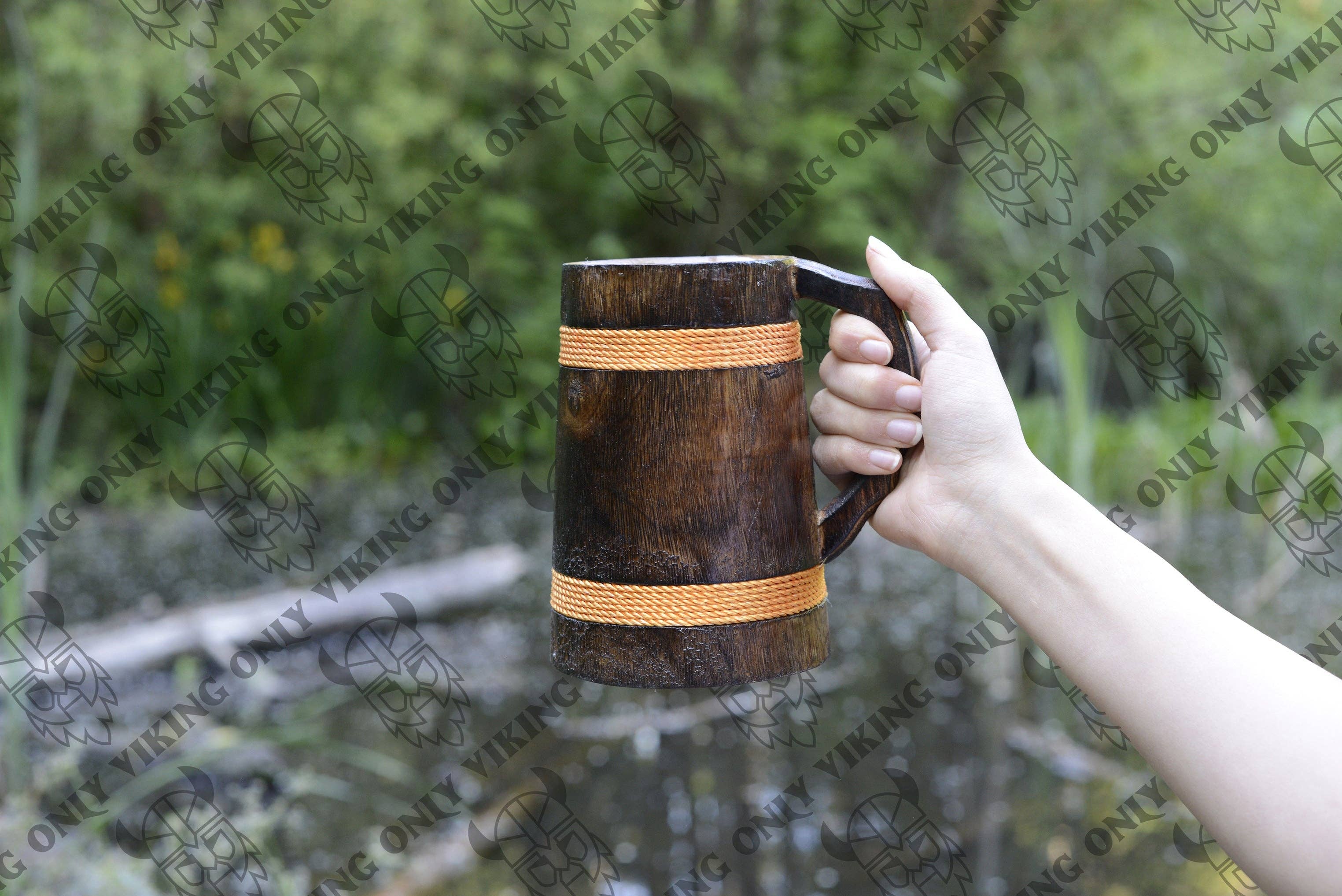 A hand holds the Mug: Wooden Viking Tankard | Vibrant Orange Hue 16-18OZ outdoors, with trees and a pond in the background.