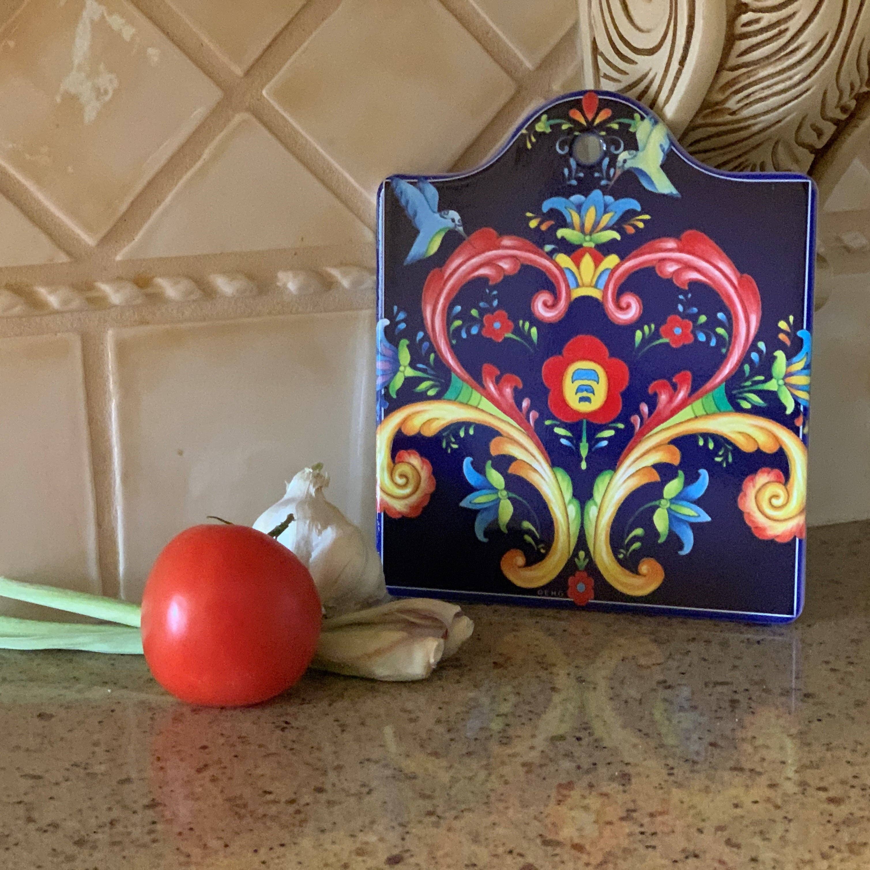 The Rosemaling Blue Ceramic Cheeseboard Kitchen Trivet, featuring a floral pattern reminiscent of Rosemaling artwork, is set on a kitchen counter with a tomato and garlic bulb nearby, against a background of tiled backsplash.