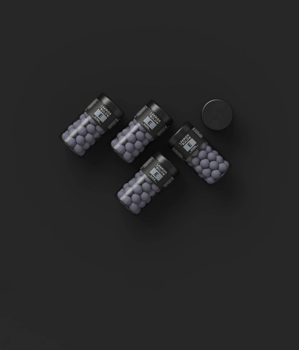Four black jars containing dark round Nordic candies, similar to salty licorice, arranged on a black background. One jar has its lid placed next to it.