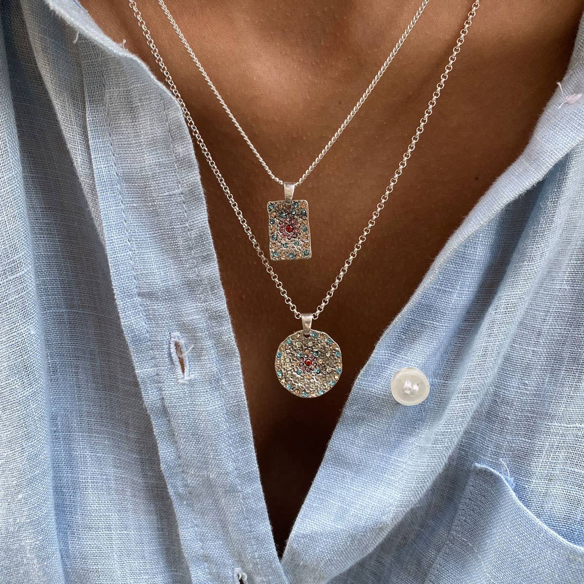 Close-up of a person in a light blue shirt wearing the A&C Oslo Vibrance Silver Pendant Necklace, featuring sparkling layered pendants—one rectangular and one circular.