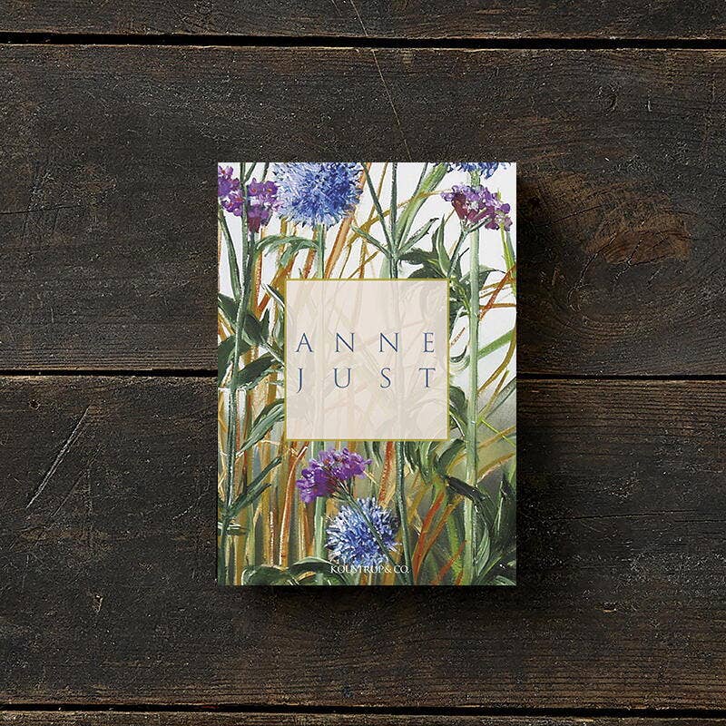 The Anne Just book lies on a wooden surface, its cover adorned with green leaves and purple flowers. Printed on FSC paper, this eco-friendly edition complements the natural elegance of the Anne Just card pack, which includes 8 cards with envelopes.