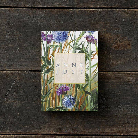 The Anne Just book lies on a wooden surface, its cover adorned with green leaves and purple flowers. Printed on FSC paper, this eco-friendly edition complements the natural elegance of the Anne Just card pack, which includes 8 cards with envelopes.