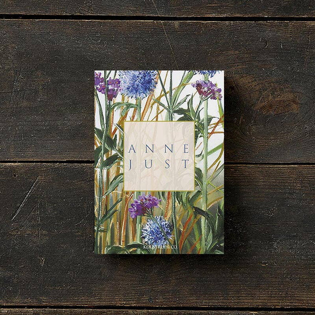 The Anne Just book lies on a wooden surface, its cover adorned with green leaves and purple flowers. Printed on FSC paper, this eco-friendly edition complements the natural elegance of the Anne Just card pack, which includes 8 cards with envelopes.