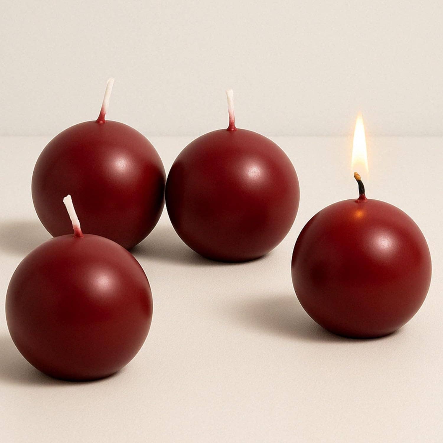 Four Candles: Burgundy Ball Candles, 2.5" Diameter, sit on a light surface; three are unscented and unlit, while one decorative candle is lit with a visible flame.