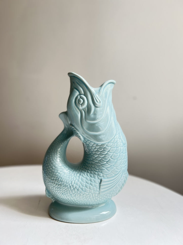 The Gluggle Jug: Eau De Nil, a light blue ceramic jug molded into a fish with an open mouth, brings an iconic touch of quirkiness to any dinner table when placed elegantly on a white surface.