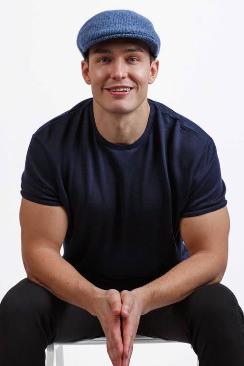 A person wearing the Hat: Wool Flat Cap: Blue and a dark t-shirt sits on a stool, smiling at the camera against a plain background.