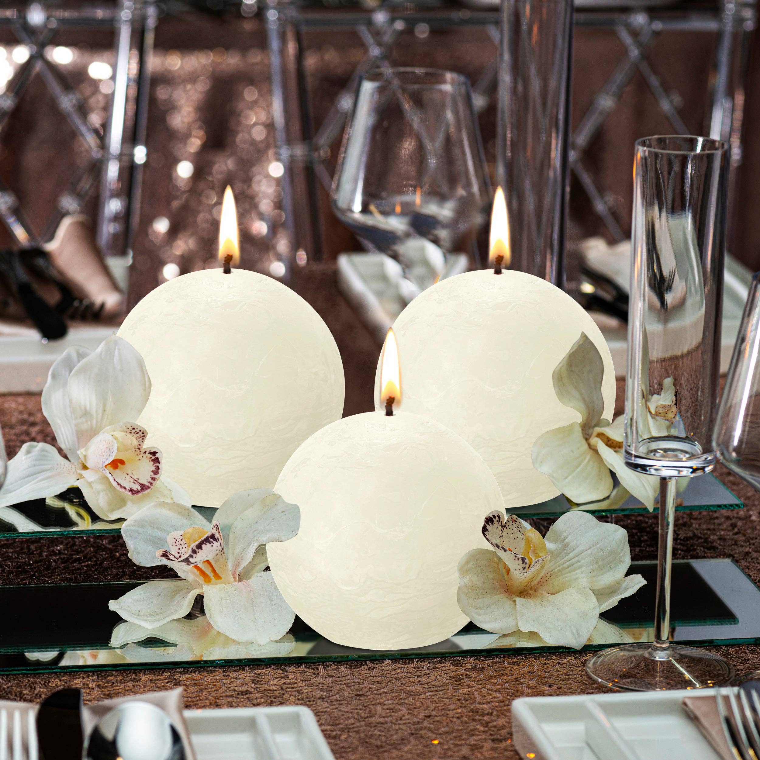 Three Blossom Pink rustic ball candles and white orchids rest on a reflective surface, surrounded by tableware, a wine glass, and a champagne flute. Made from eco-friendly plant-based wax, these unscented beauties enhance any setting with elegance.