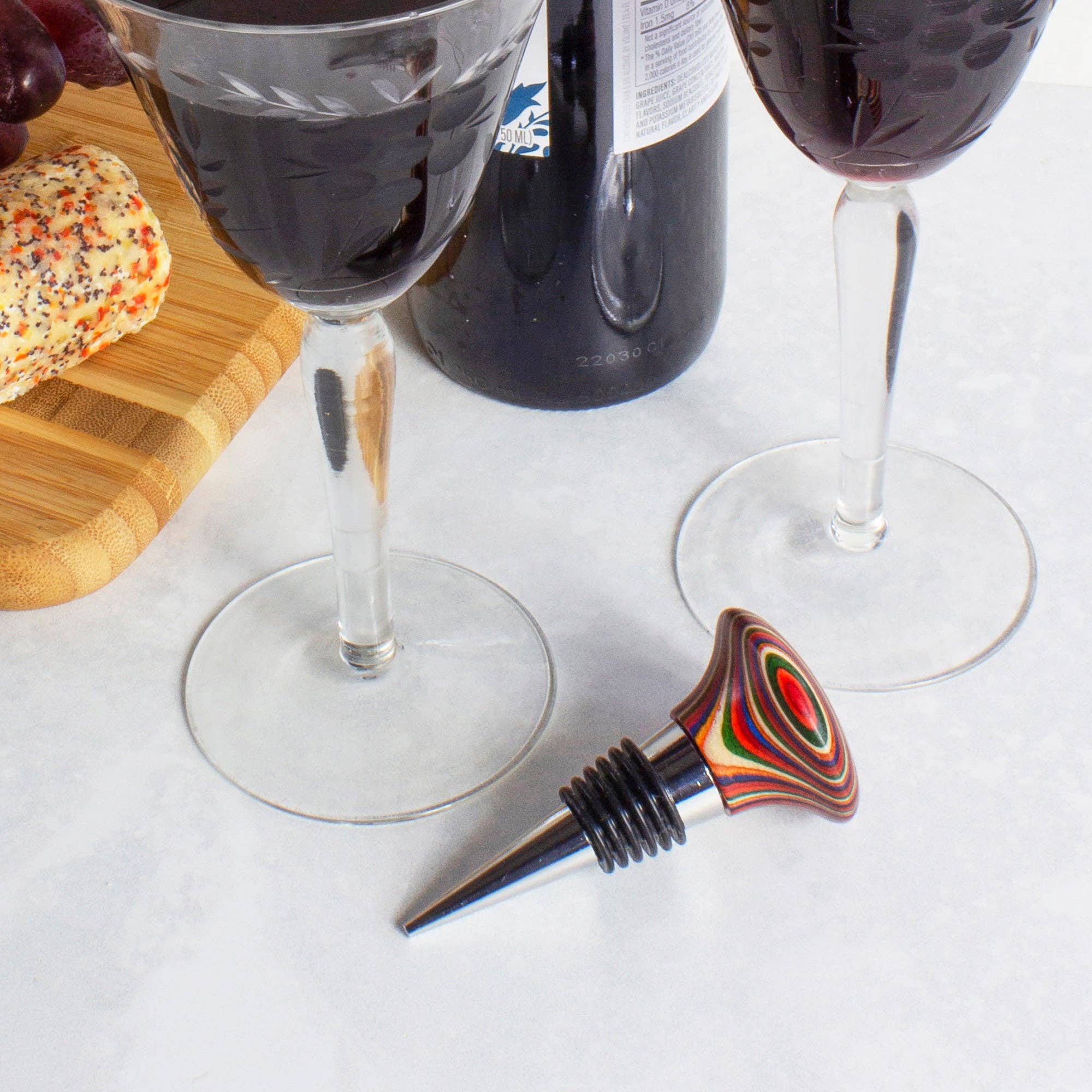 Two glasses of red wine sit beside a colorful Marrakesh Collection Wine Bottle Stopper and wine bottle on a table with a cheese board, enhanced by air-tight silicone rings to preserve freshness.