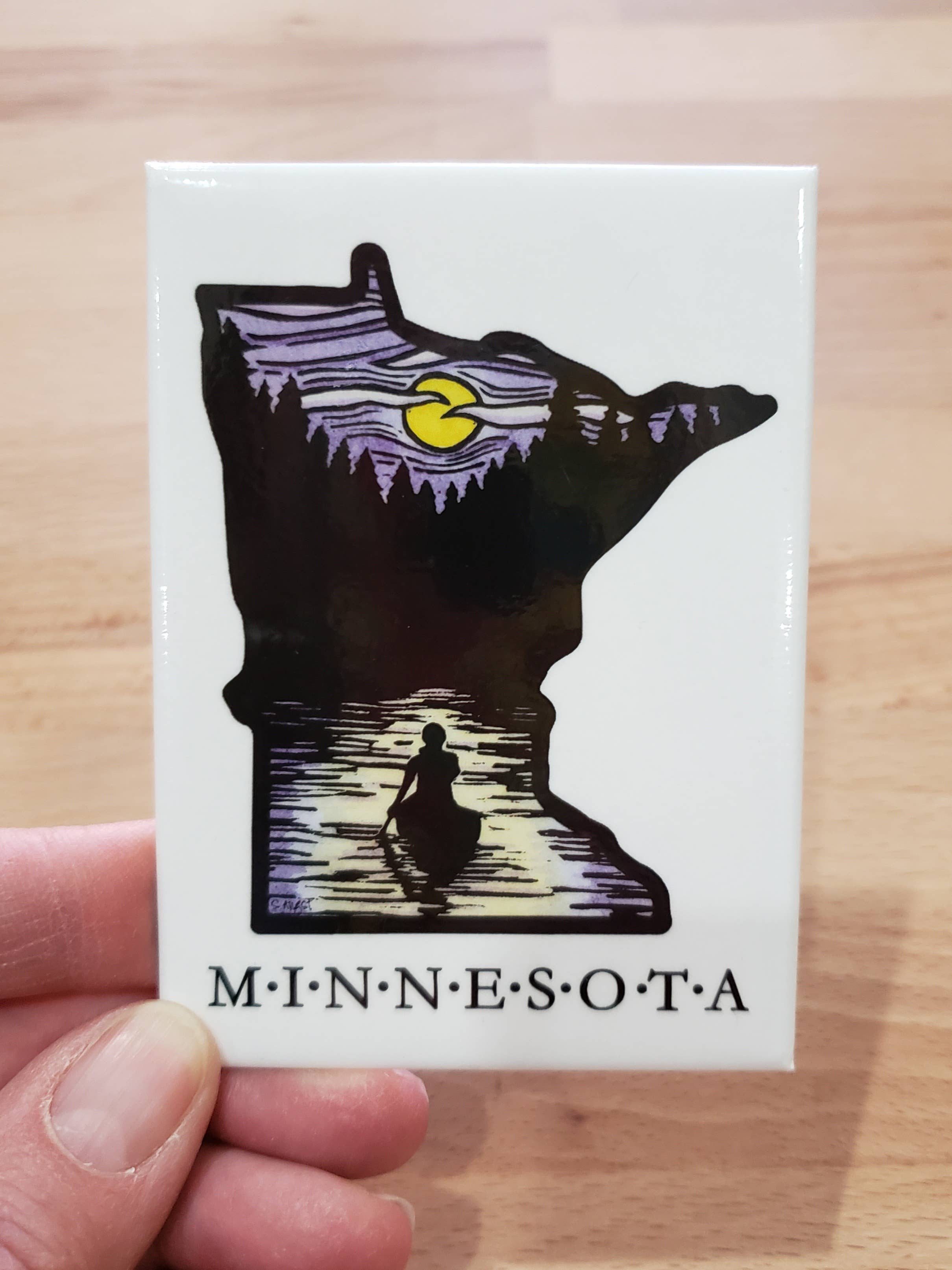 The Magnet - Minnesota Solitude Canoe Magnet features a strong magnet shaped like Minnesota with a silhouette of a person in a canoe on a calm lake, framed by moonlit skies and a forest background.