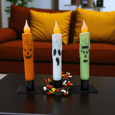 Three Frankenstein Halloween Battery Operated Timer Taper Candles with jack-o-lantern and ghost designs are arranged on a black tray in front of an orange sofa, bringing a festive touch to your Halloween décor.