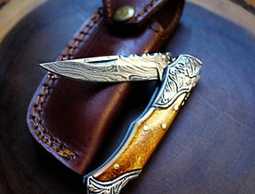The Titan International Damascus Steel handmade pocket knife with burnt bone handles features an etched blade and sits atop a brown leather belt sheath with visible stitching.