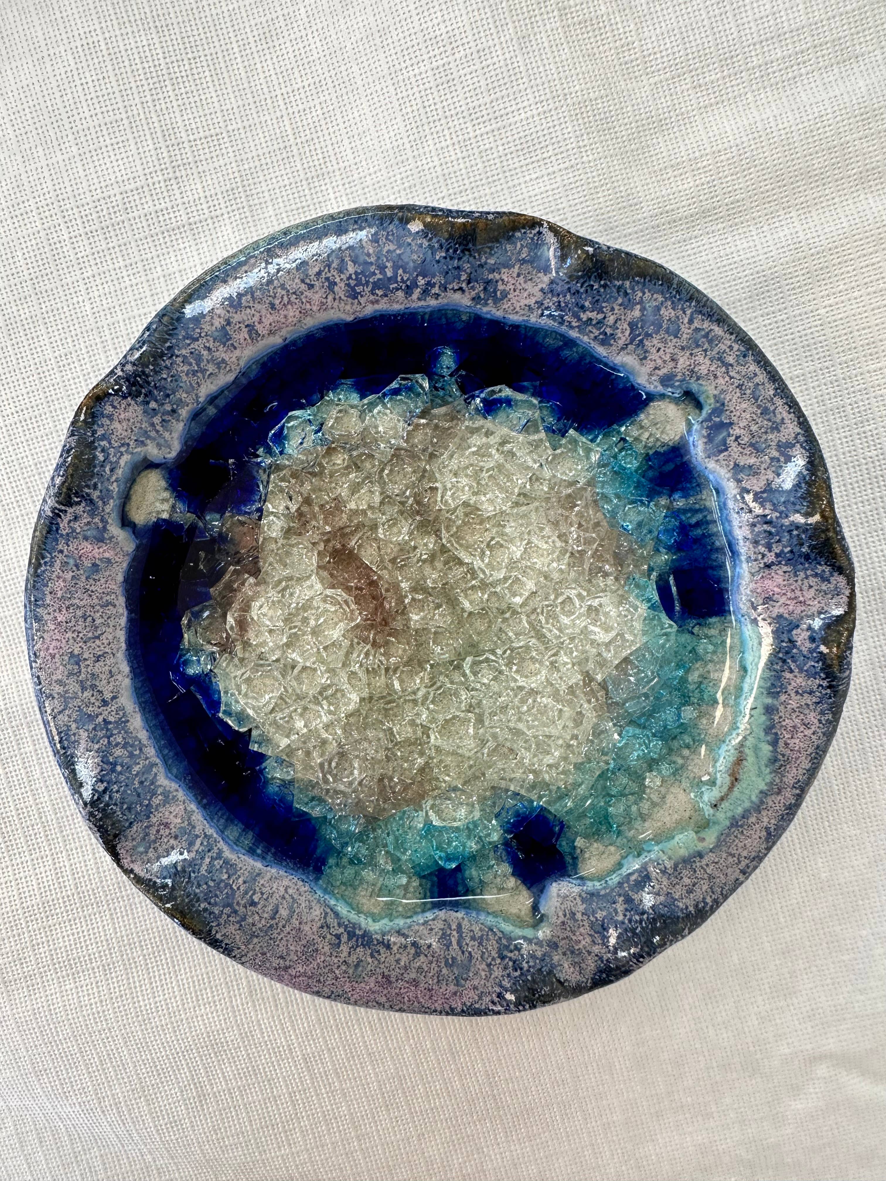 A round, glazed ceramic candle holder, inspired by handmade pottery, features a textured, crystalline interior. The glaze shows shades of blue and green with a mottled rim. It rests on a light fabric surface, embodying the charm of artisanal craft.