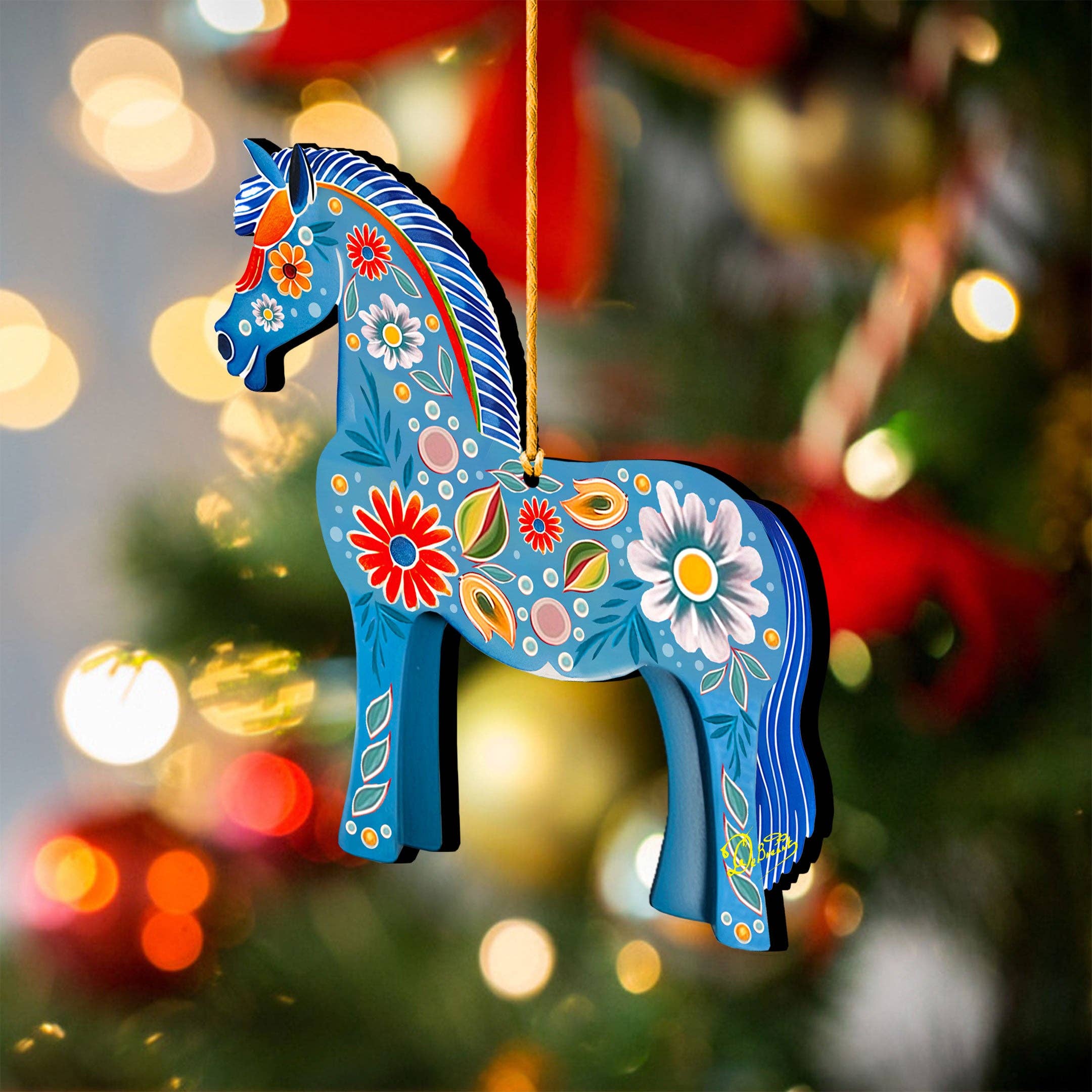 A beautifully handcrafted wooden ornament, named the Danish Horse Decorative Wood Ornaments GDebrekht Love Family, with floral patterns and a decorative blue design reminiscent of Danish Horse Ornaments, elegantly hangs on a Christmas tree amidst softly blurred lights in the background. This piece showcases the exquisite artistry of G.DeBrekht Art.