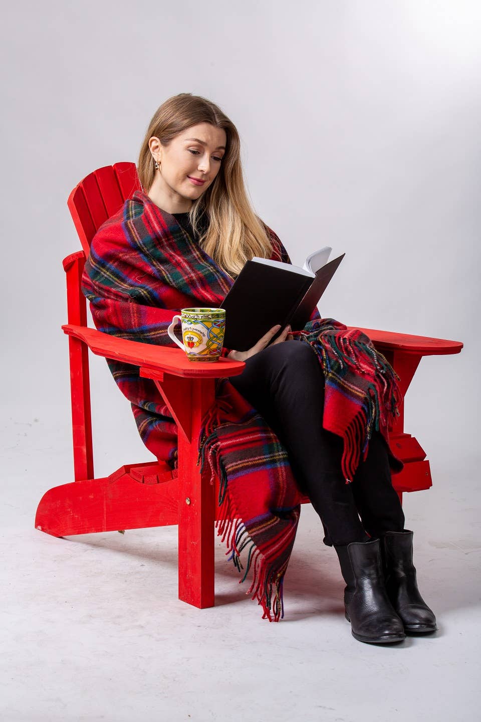 A woman sits in a red chair, wrapped in a Royal Stewart Deluxe Highland Blanket, reading a book with a colorful mug beside her.