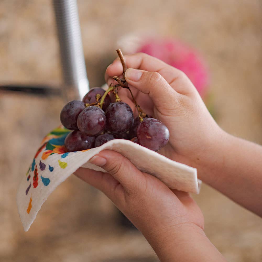 Hands hold a bunch of red grapes over a vibrant, sustainable Home Sweet Home Swedish Dishcloth.