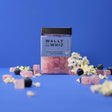 A jar of Wally & Whiz - Elderflower with Blueberry Gummy Cubes (240g) Danish candy sits on a blue surface, surrounded by gummies, elderflowers, and blueberries against a blue background.
