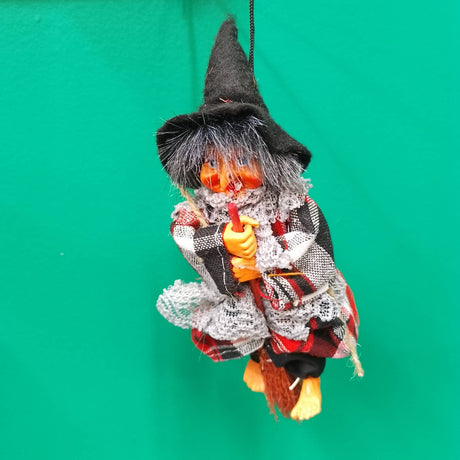 The Kitchen Witch: Minna Red Doll, featuring gray hair, a black hat, and a plaid outfit, sits on a broomstick, creating delightful folklore décor against a bright green background.