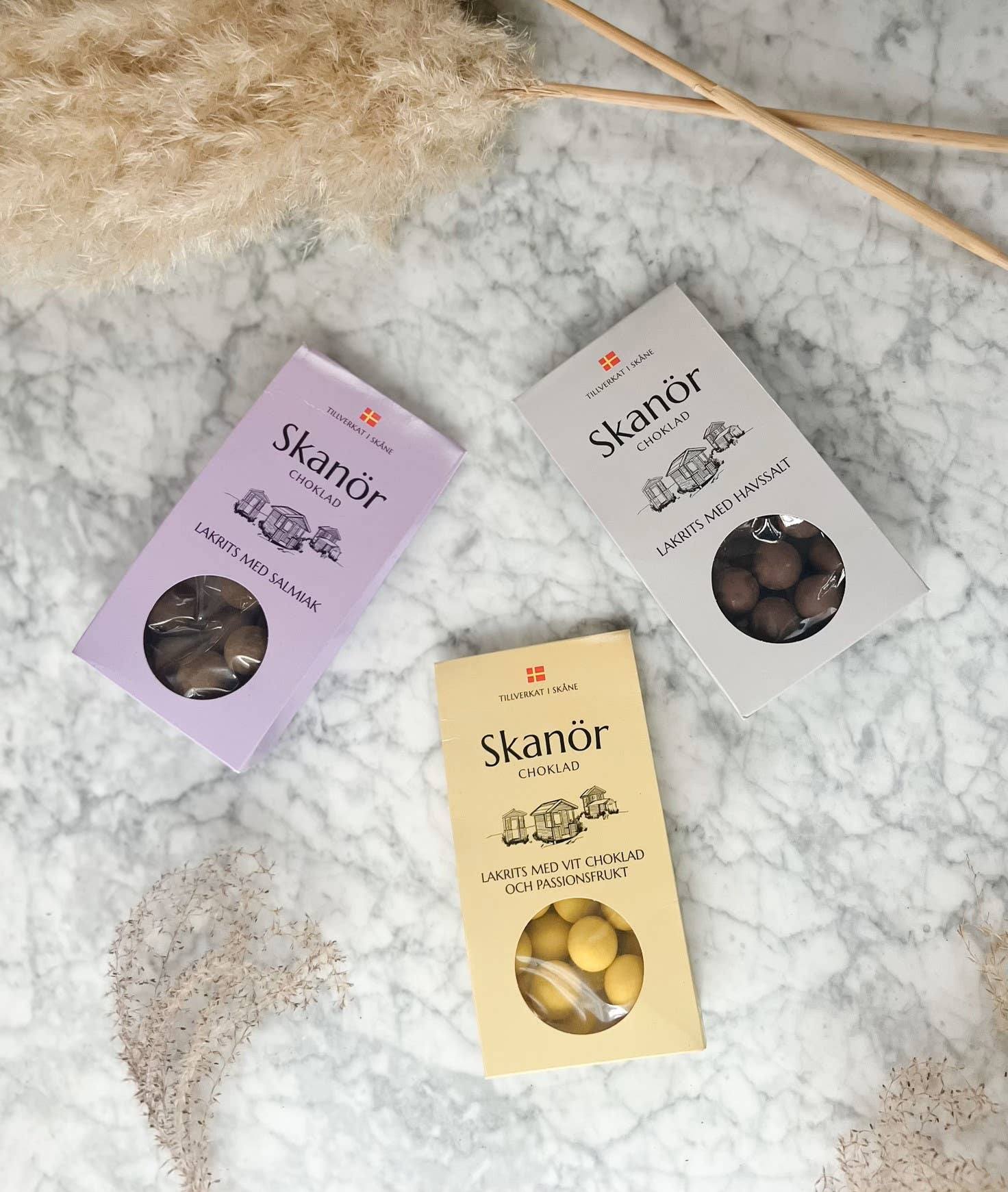 Three vibrant boxes of Skanör chocolates—including exquisite flavors like licorice with sea salt, hazelnut, and passion fruit—are beautifully displayed on a marble surface alongside dried pampas grass. Crafted in Skåne, Sweden, these delectable treats offer a delightful journey of unique flavors with their smooth milk chocolate essence.