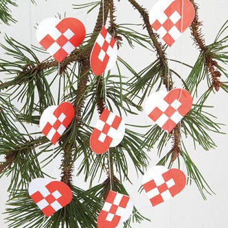 The Ornament: Braided Heart Garland in Danish Flag Colors Red & White hangs gracefully from pine tree branches, featuring several red and white hearts inspired by the Danish flag.