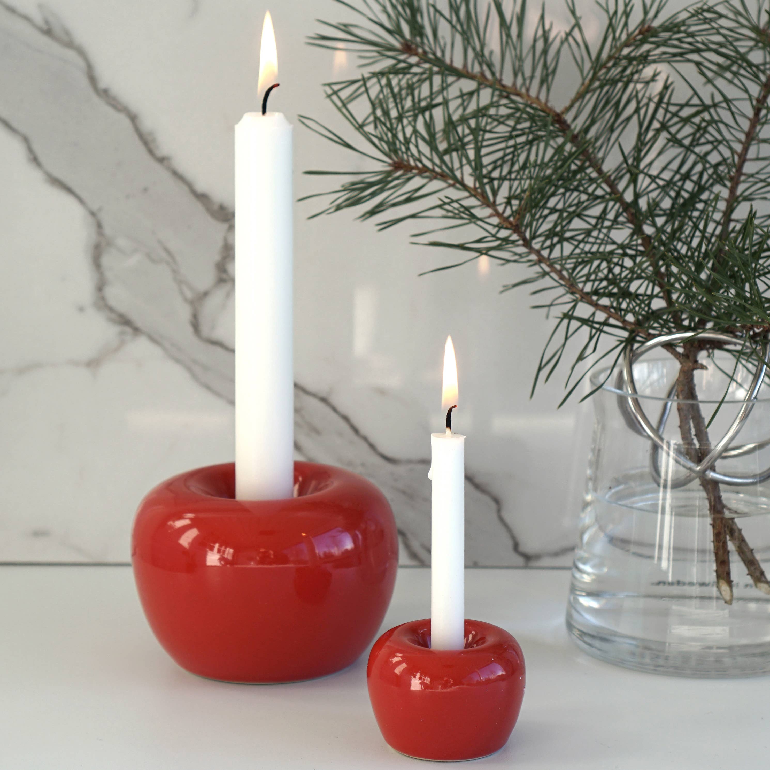 Two lit white candles stand elegantly in Swedish shiny red apple taper candle holders, accompanied by a small branch with green needles in a clear glass vase. The scene is set against a white marble surface with subtle gray veins, enhancing the elegant and serene atmosphere.