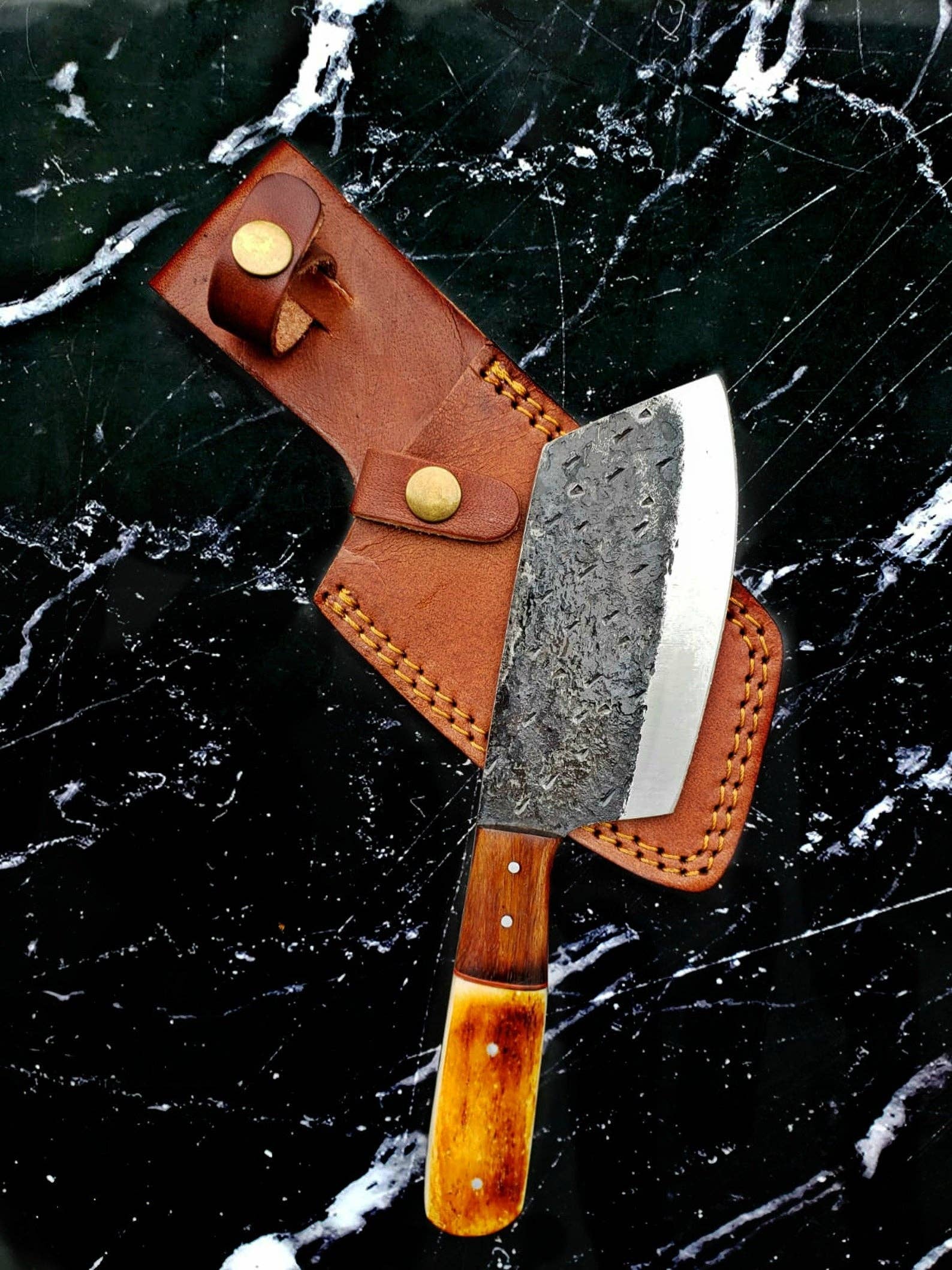 The Knife: Handmade Carbon Steel Cleaver for Kitchen & Outdoor Use features a rustic wooden handle and rests in a brown leather sheath on black marble, highlighting its robust, handcrafted design.
