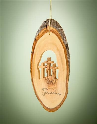 The Ornament: Bark Slice with Three Crosses - 4 to 6", hung by a string against a green background, evokes the spirit of the Holy Land with its olive wood design and engraved "Jerusalem".