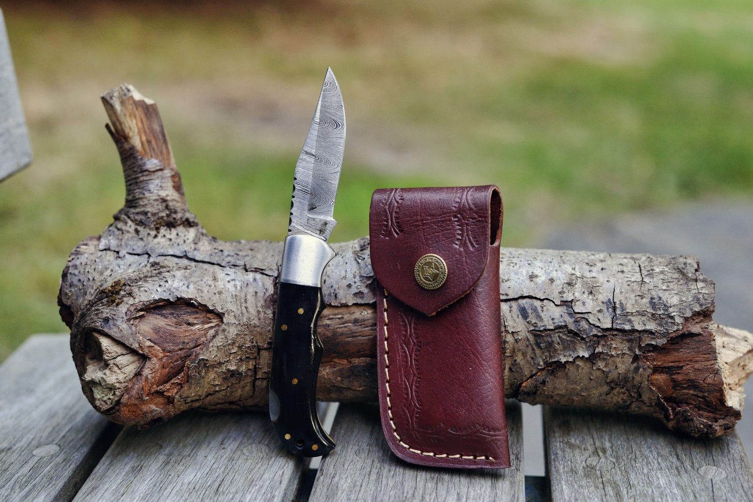 The Buffalo Horn Genuine Damascus Folding Knife, featuring a sleek black handle and an intricately engraved blade, is elegantly displayed on a wooden surface. It rests on a log alongside its brown leather sheath, presenting an ideal gift for men.