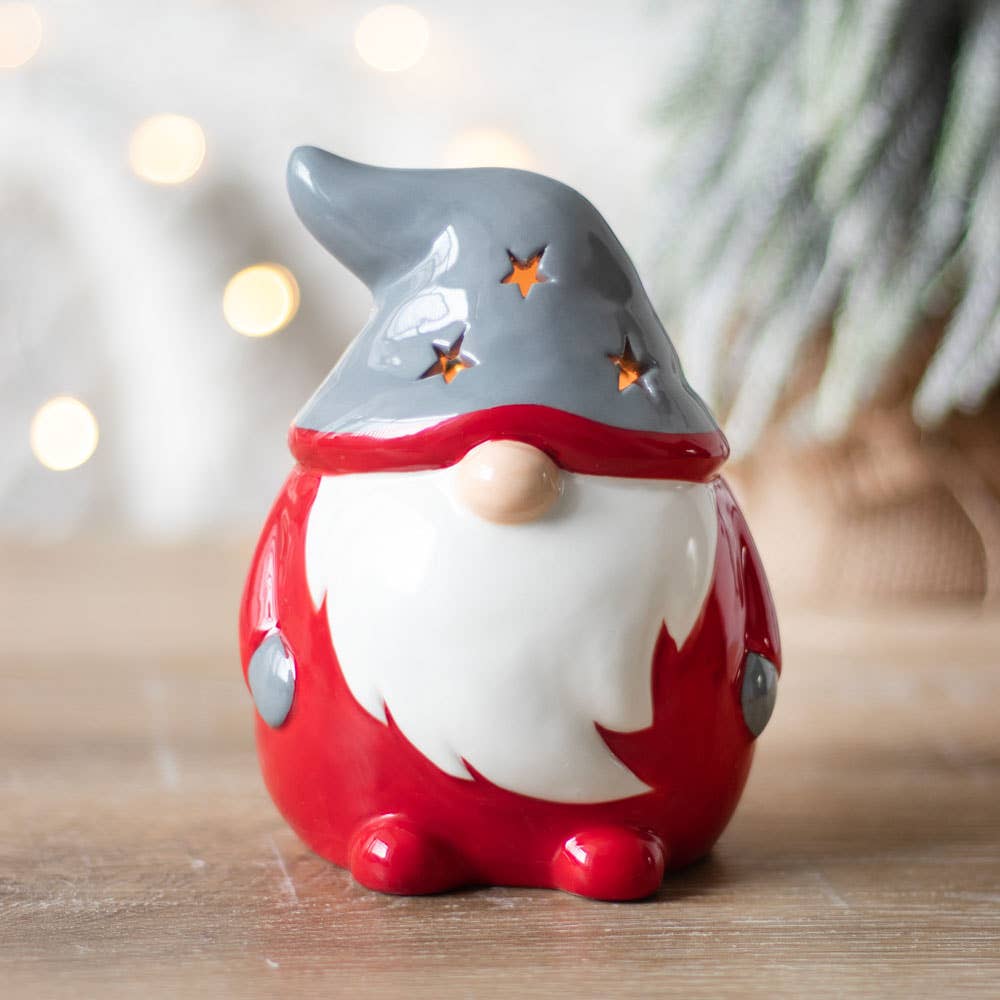 The Candle Holder: Gnome Tealight Holder showcases a ceramic gnome figurine dressed in a red outfit and gray hat with star cutouts, daintily placed on a wooden surface. Its delightful charm is amplified by the gentle glow of blurred lights in the background, serving as both a decorative piece and a tealight holder for an extra touch of warmth and cheer.