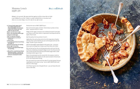 A copy of "Scandinavian Year" by Bronte Aurell lies on a blue surface next to a partially eaten apple pie, its filling exposed where a slice is missing, while the pie dish and serving knife rest among scattered crumbs.