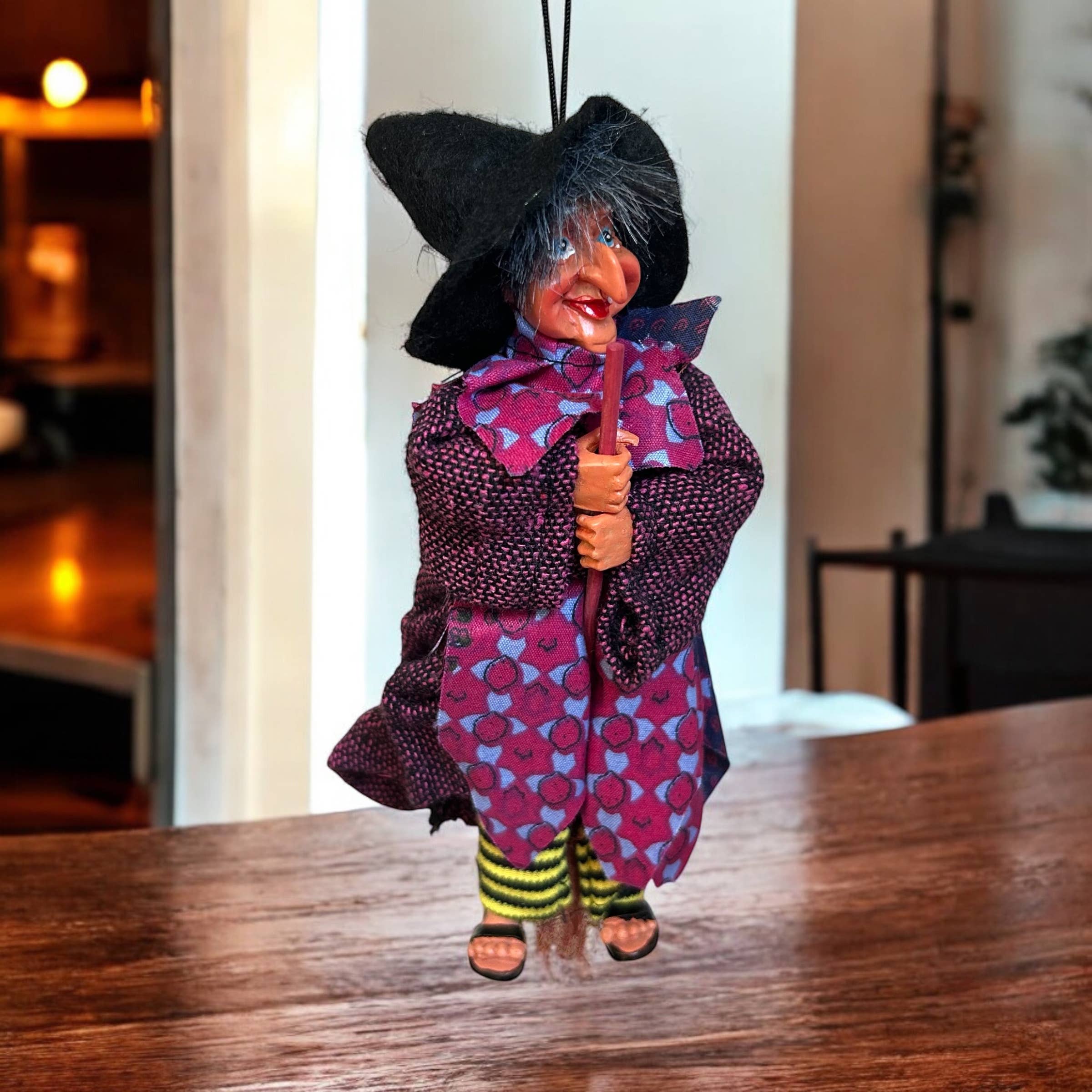 The Figurine: Johanna Purple Kitchen Witch Doll, 9" tall, wears a black hat, purple dress, striped socks, and sandals. Hanging by a string and holding a broomstick, she makes a charming good luck charm for your home.