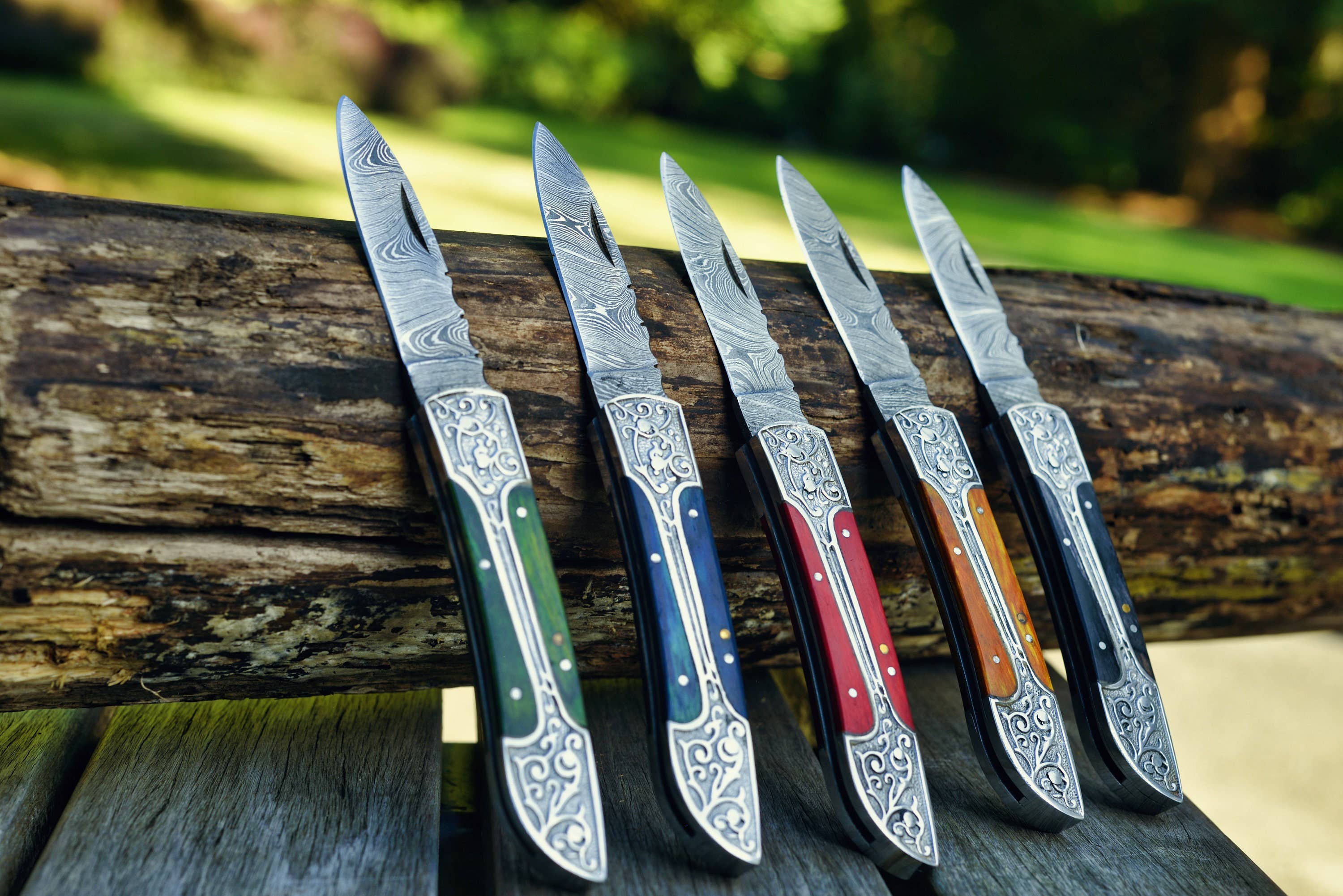 Five Black Damascus folding pocket knives with intricate designs are elegantly displayed against a rustic wooden log outdoors, making them ideal gifts for men.