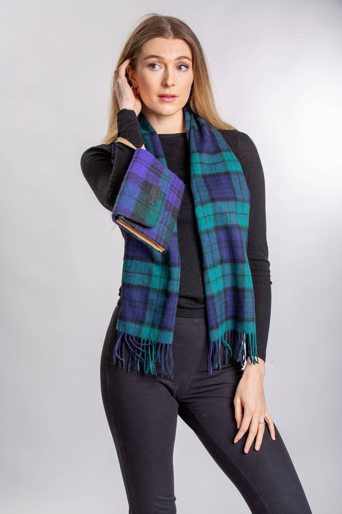 A woman in a long-sleeved black top and pants models the Blackwatch Lambswool Scarf by Patrick King, featuring a blue and green plaid pattern, against a neutral background.