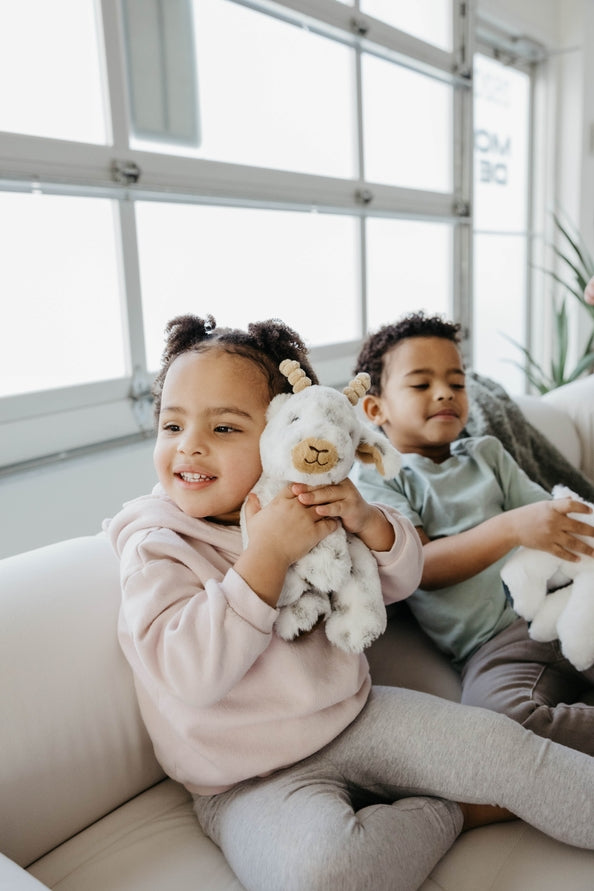 Two young children sit on a white couch indoors, smiling and holding plush stuffed animals. The girl in front hugs the Faire - The Petting Zoo Plush: Goat 9″ Tall Stuffed Animal, while the boy in the background holds a white stuffed animal.