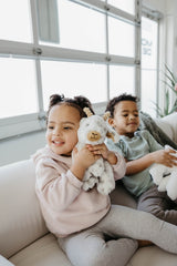 Two young children sit on a white couch indoors, smiling and holding plush stuffed animals. The girl in front hugs the Faire - The Petting Zoo Plush: Goat 9″ Tall Stuffed Animal, while the boy in the background holds a white stuffed animal.
