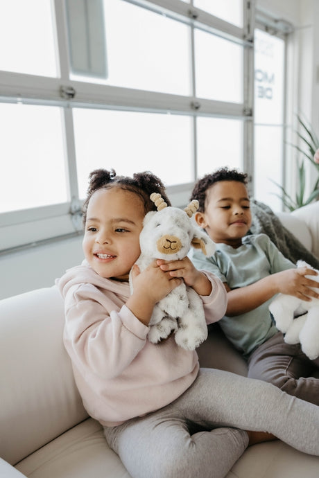 Two young children sit on a white couch indoors, smiling and holding plush stuffed animals. The girl in front hugs the Faire - The Petting Zoo Plush: Goat 9″ Tall Stuffed Animal, while the boy in the background holds a white stuffed animal.